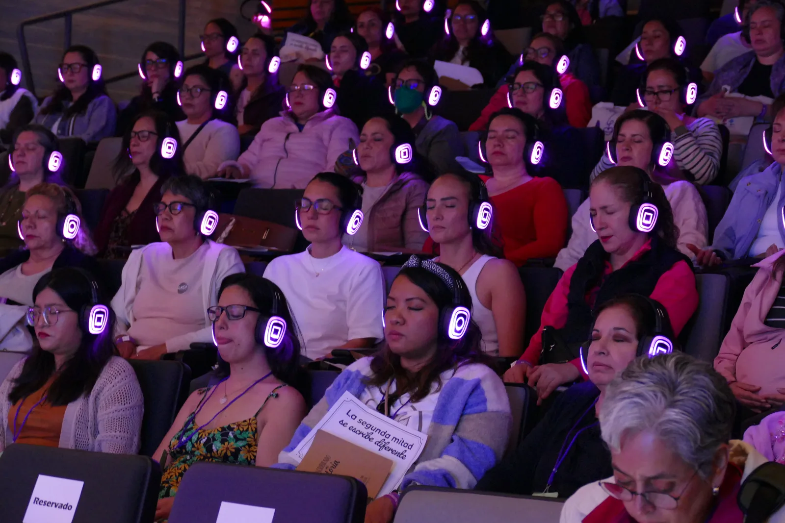 Asistentes con audífonos luminosos, meditando con los ojos cerrados en el auditorio.